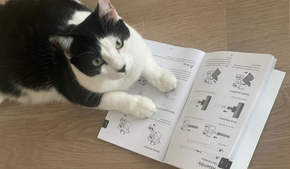 A black and white cat is shown siting on a vacuum instruction manual for Yahoo Life's Best Cordless Stick Vacuum guide.