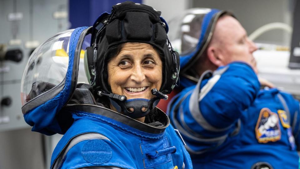 two astronauts in spacesuits, with suni williams in the front turning to smile at the camera