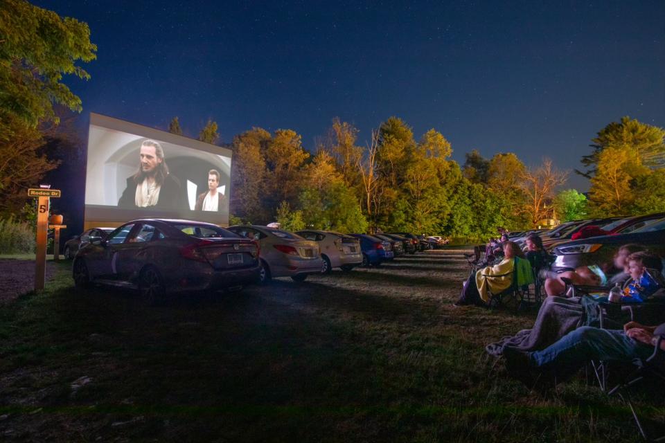 Cars lined up and people sitting outside in folding chairs at Four Brothers Drive-In during an evening showing.