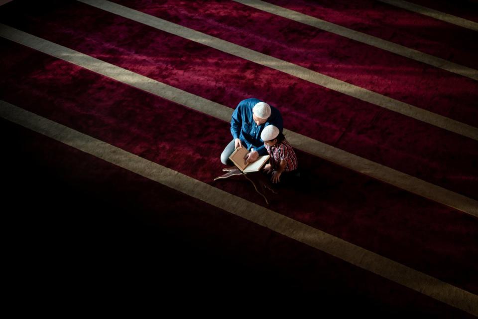 A man and a child, both wearing white head coverings, kneel as they read a book on a huge red carpet.