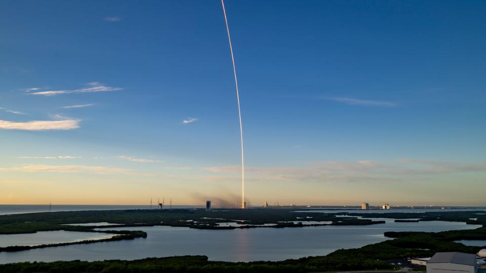 a bright streak of light arcs from the ground into the sky above a marshy wetland area in the early morning