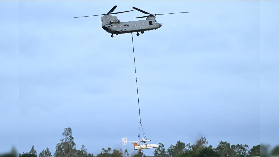 a large twin-rotor helicopter carries a white windowless aircraft beneath it at the end of a long cable