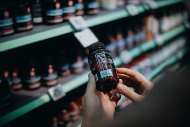 <p>d3sign / Getty Images</p> Person reading the label on a bottle of medicine at a pharmacy.