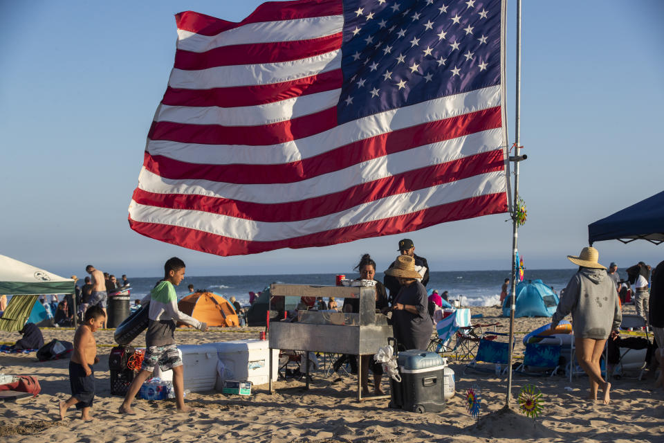 Huntington Beach, CA - July 04: The Espinoza, Vazquez and Romero families of Fallbrook grill tacos on the beach beneath their giant American flag as they await the fireworks over the ocean Sunday, July 4, 2021 in Huntington Beach, CA. The festival featured live entertainment & DJs, merchant & crafts vendor booths, amusements & carnival rides, food trucks, beer & wine garden, sponsor activations, live radio broadcasts, and prize giveaways. A Surf City Run 5K kicked off the day and a neighborhood parade that included 3 separate routes replaced the large parade of years past before the pandemic. The grand finale featured fireworks over the ocean shot off from the pier at 9pm. (Allen J. Schaben / Los Angeles Times via Getty Images)