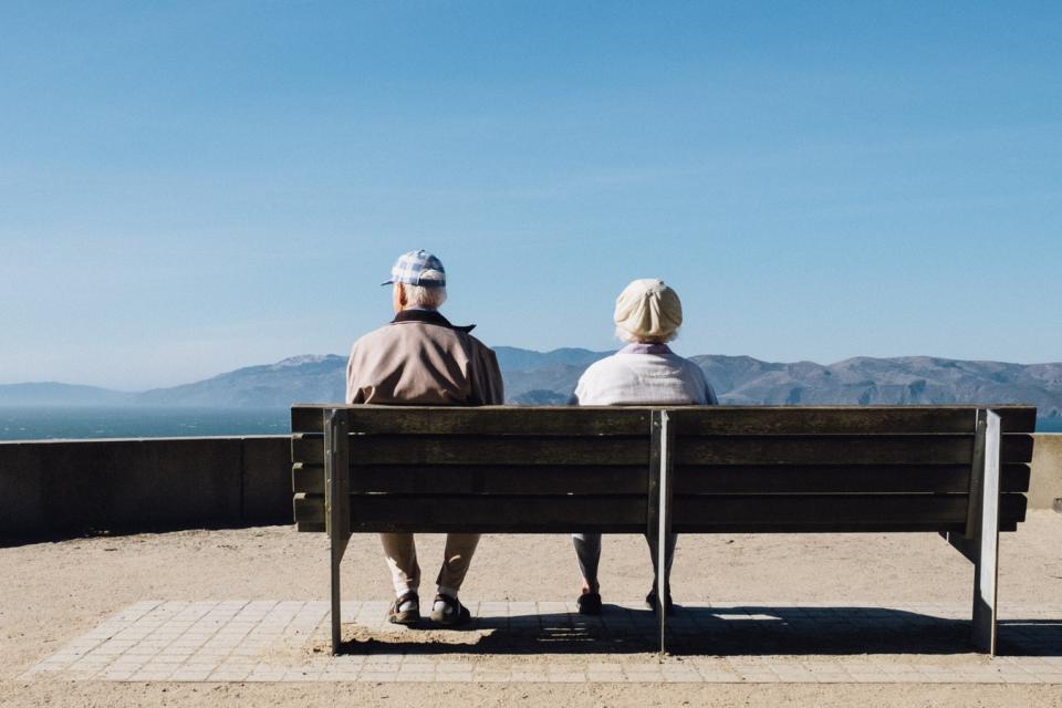 An older man and woman sitting on a park bench looking out at a body of water.