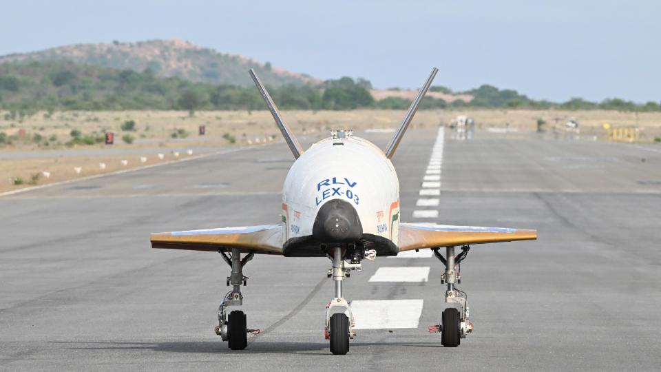 a white aircraft with no windows or cockpit canopy lands on a runway surrounded by tropical vegetation