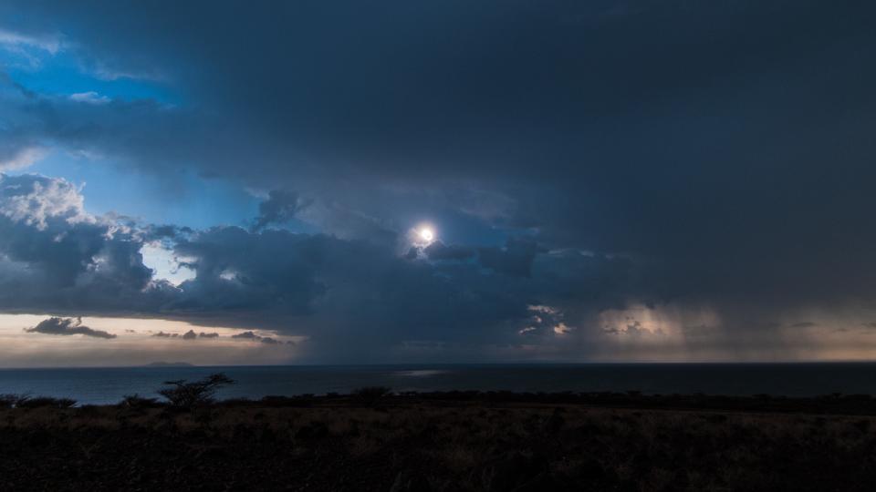 eclipsed sun shines behind many storm clouds with rain visible in the distance.