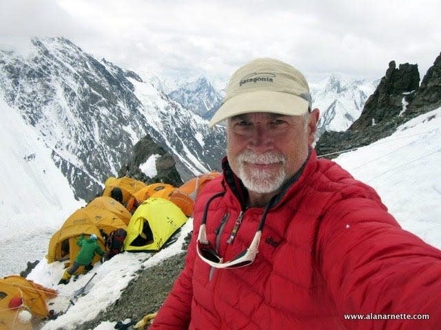 A selfie taken by a man wearing a red coat and beige Patagonia hat standing above a snowy camp on K2