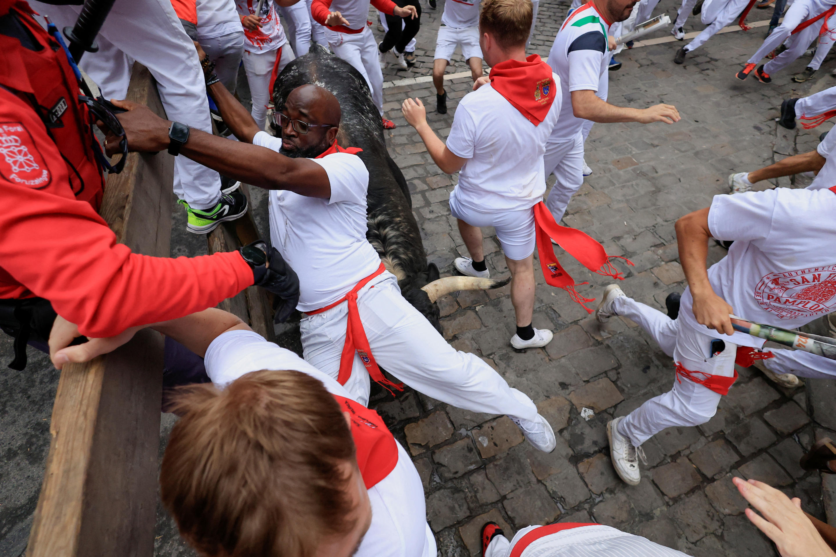 A reveler dressed in white with a red scarf hangs on to a wooden structure as a bull runs down the street, seemingly right beneath him.