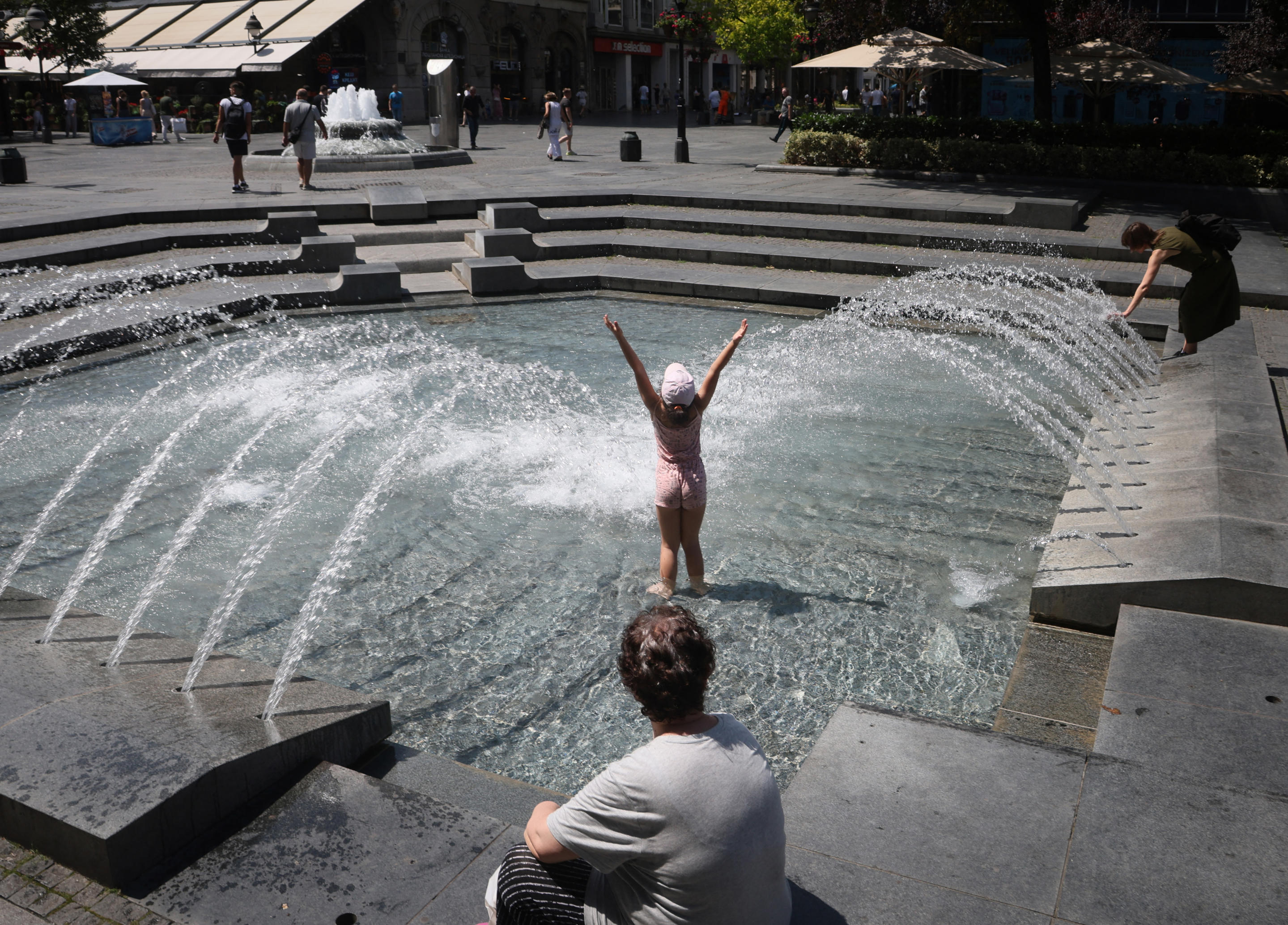 A young girl wearing a bathing suit with her arms outstretched high in the air plays in a public water fountain.