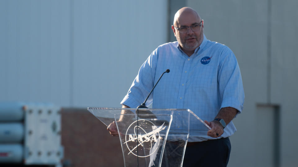 A bald man in a collared shirt with a nasa logo stands a a clear podium with a nasa logo, in front of a hangar in the background.