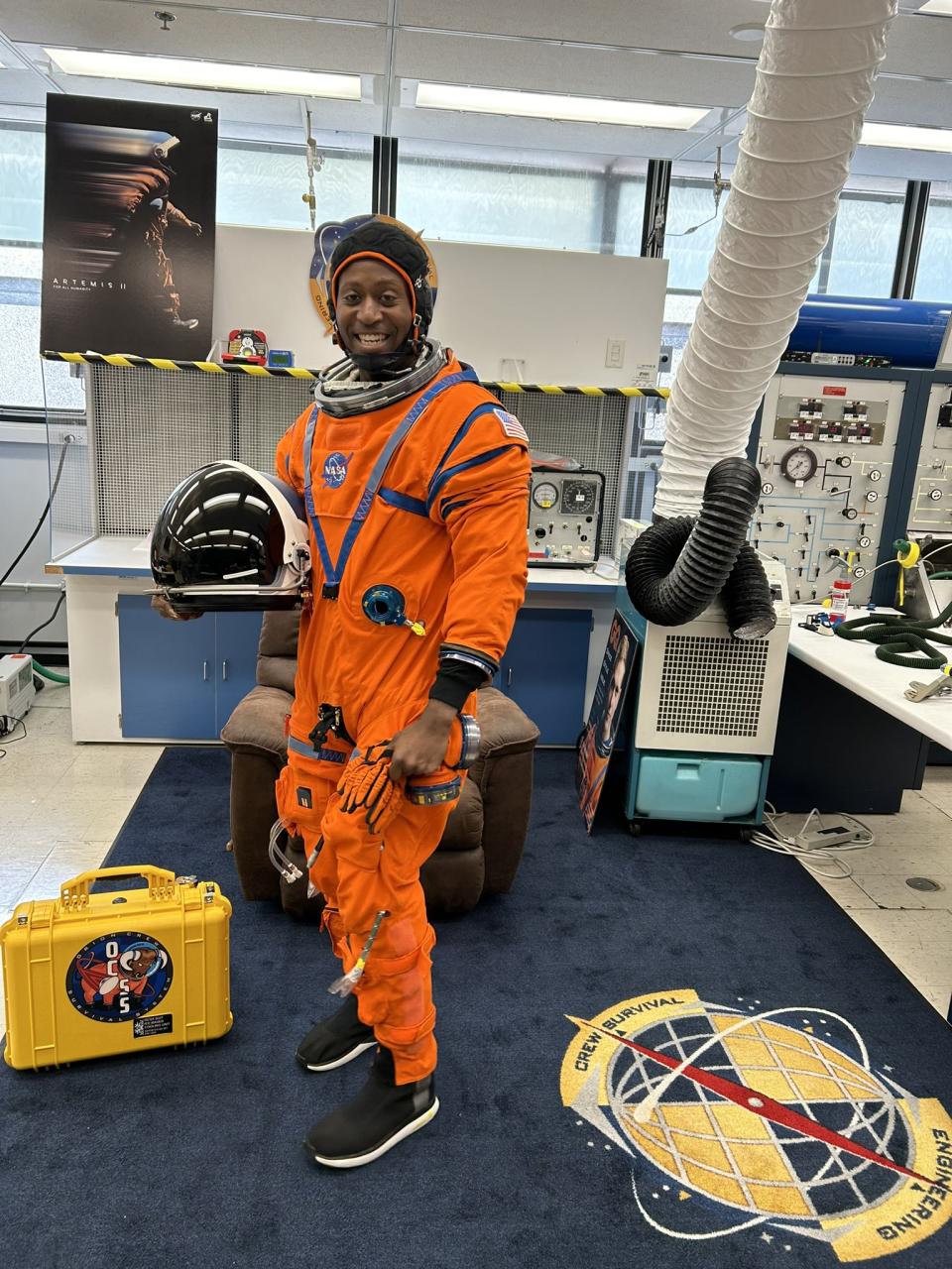 astronaut andre douglas smiling while standing up in an orange spacesuit, in an office filled with moon posters