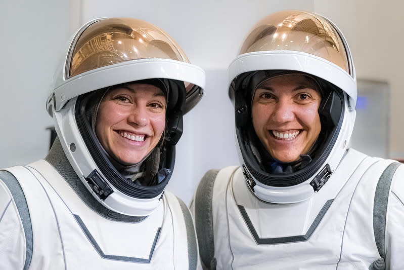 Two women in white space suits are seen from the chest up. Their copper colored visors are flipped up to show their faces cushined inside their helmets. They are in a white room smiling.