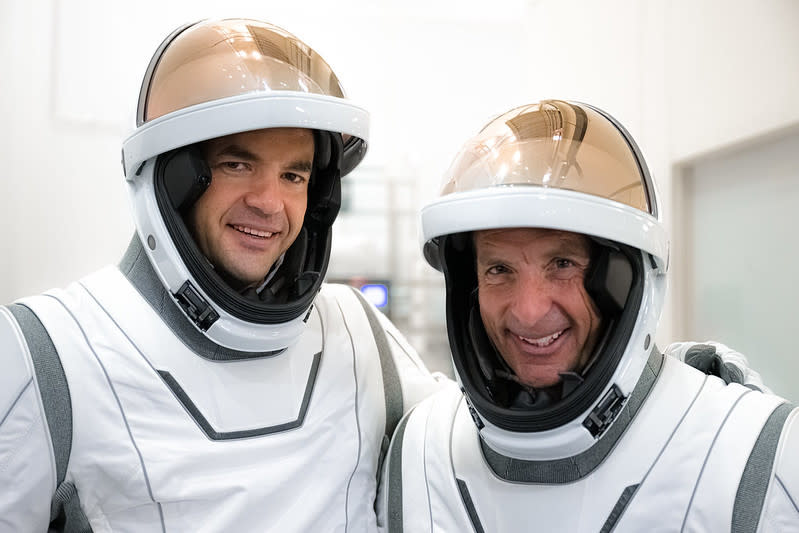 Two men in white space suits are seen from the chest up. Their copper colored visors are flipped up to show their faces cushined inside their helmets. They are in a white room smiling.