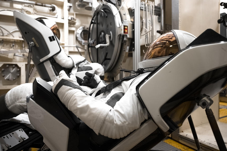 Two astronauts, one in the foreground facing left, the other in the background facing right, lean back in white and black bucket sets, wearing white spacesuits.