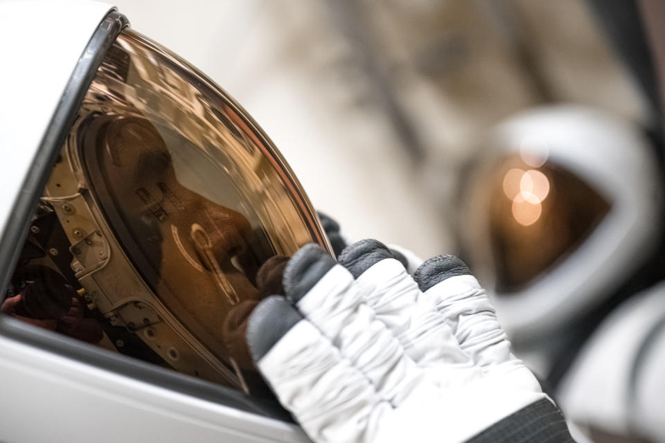 A closeup of a white-shelled astronaut helmet from the left, extending to the middle of the image. A copper-shaded visor stretches across the dimmed interior face. A black-tipped white glove tests on the visor.