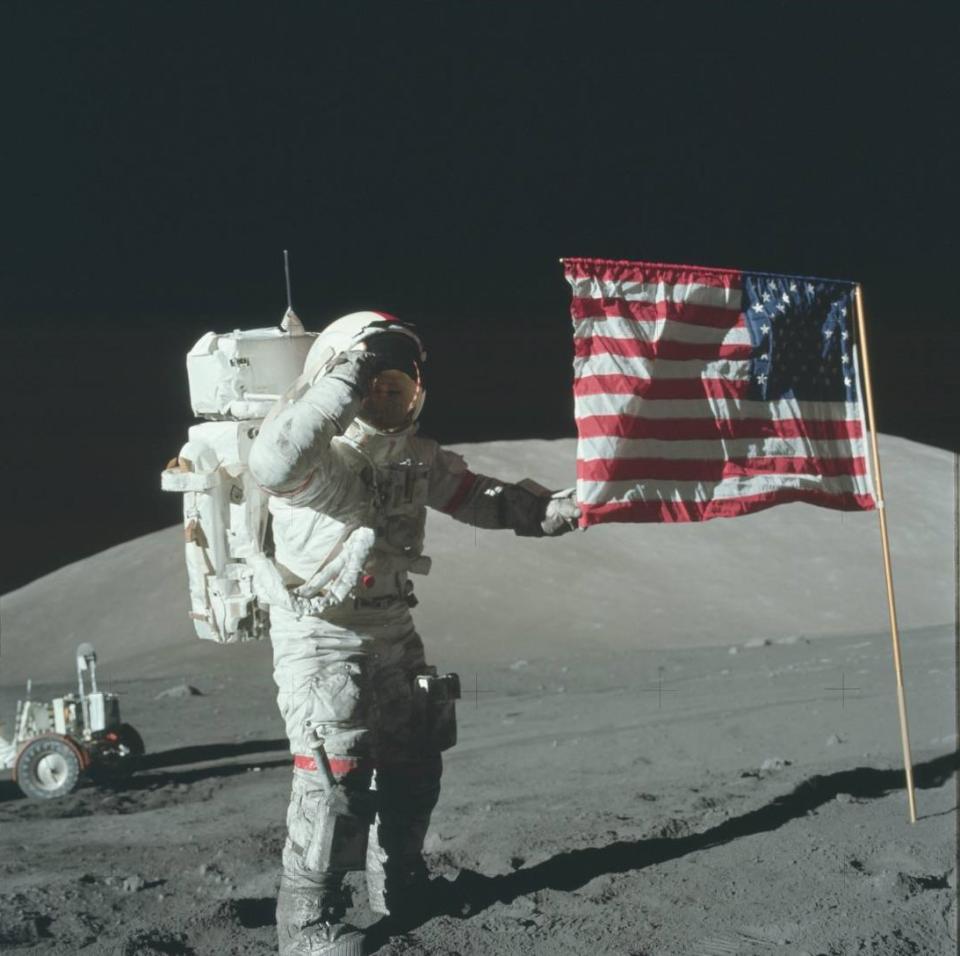 an astronaut in a bulky white spacesuit plants an american flag on the dusty grey surface of the moon