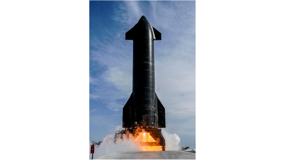 a large black rocket fires its engines on an outdoor test stand, with blue sky in the background