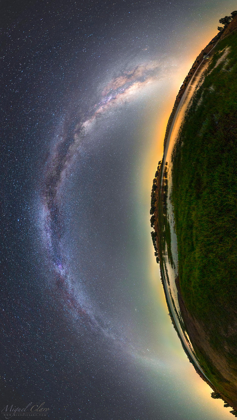 an arc of hazy white light stretches across a starry night sky next to a curved panoramic view of a lush green landscape. the whole image is rotated 90 degrees counterclockwise so that the sky is on the left and the landscape is on the right. the combination of the arc of white light and the arc of the ground forms almost an eye-like shape