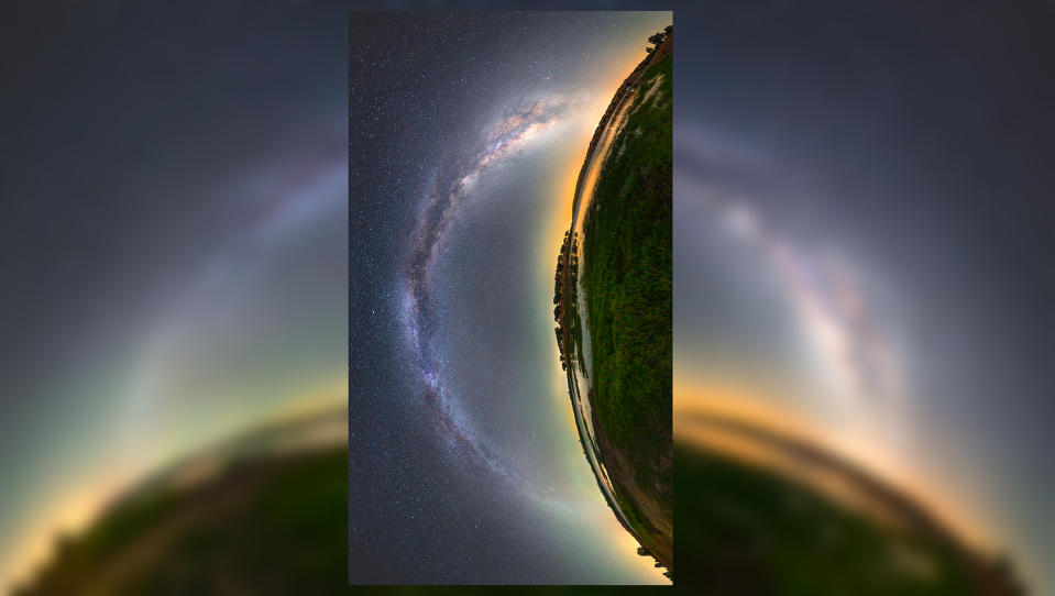  An arc of hazy white light stretches across a starry night sky next to a curved panoramic view of a lush green landscape. the whole image is rotated 90 degrees counterclockwise so that the sky is on the left and the landscape is on the right. the combination of the arc of white light and the arc of the ground forms almost an eye-like shape. 