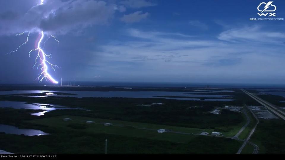  A bright white lightning bolt strikes a launch pad in florida, seen from a distance with water and greenery in the foreground. 