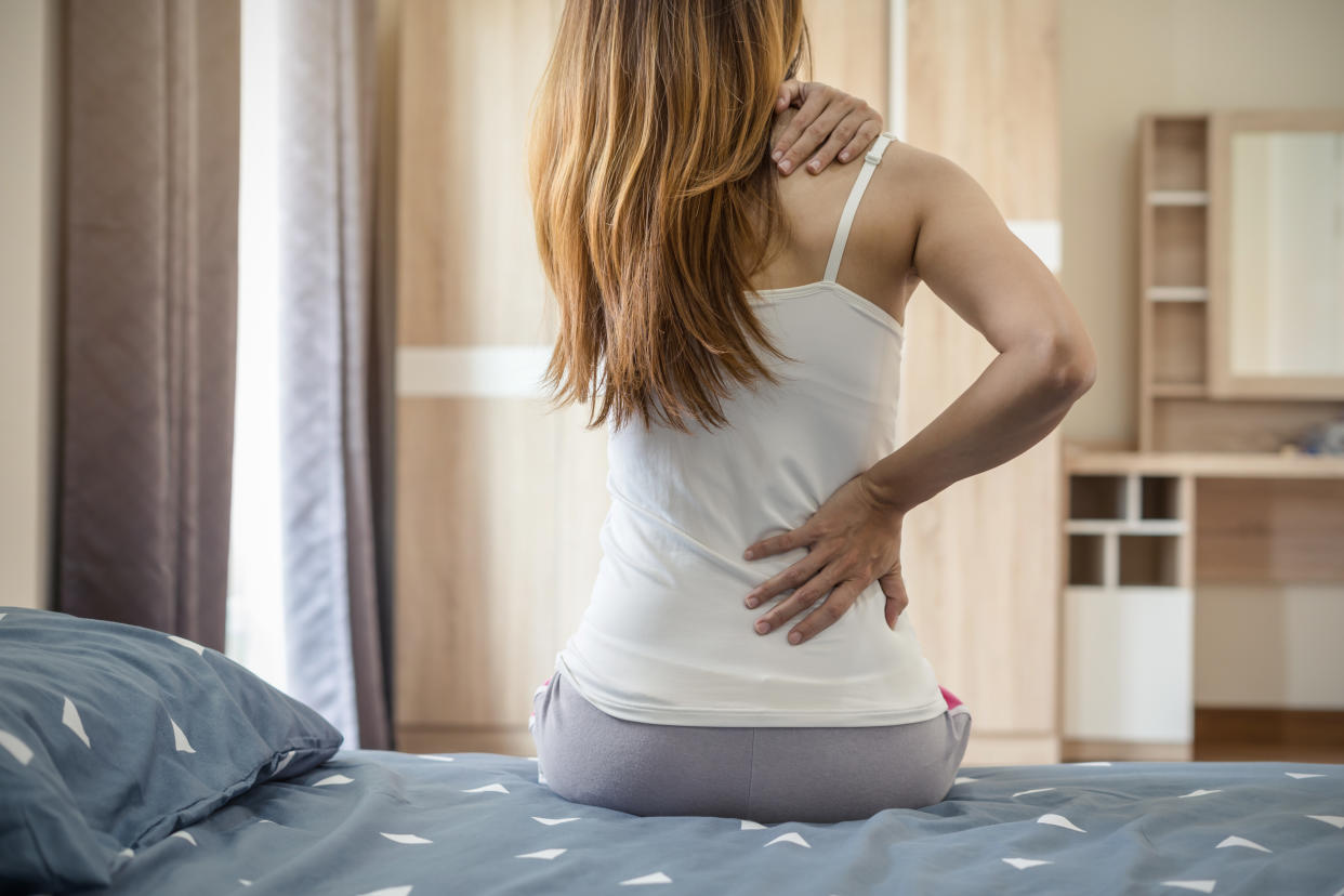 A white woman with brown hair sitting on a modern-looking bed holds her lower back and right shoulder 