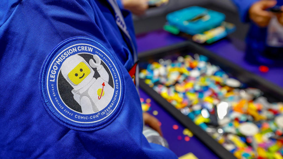  Closeup of a patch on a blue shirt, which shows a spacesuited lego man smiling and waving. blurry bins of legos are in the background. 