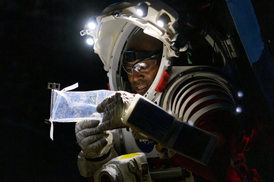 an astronaut in a spacesuit at night. he looks closely at a plastic bag that has a sample inside it