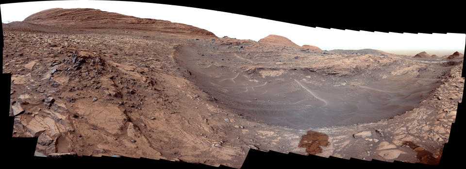 A reddish landscape with a mountain in the background and rocky areas in the foreground. A large, shallow valley is seen toward the right.