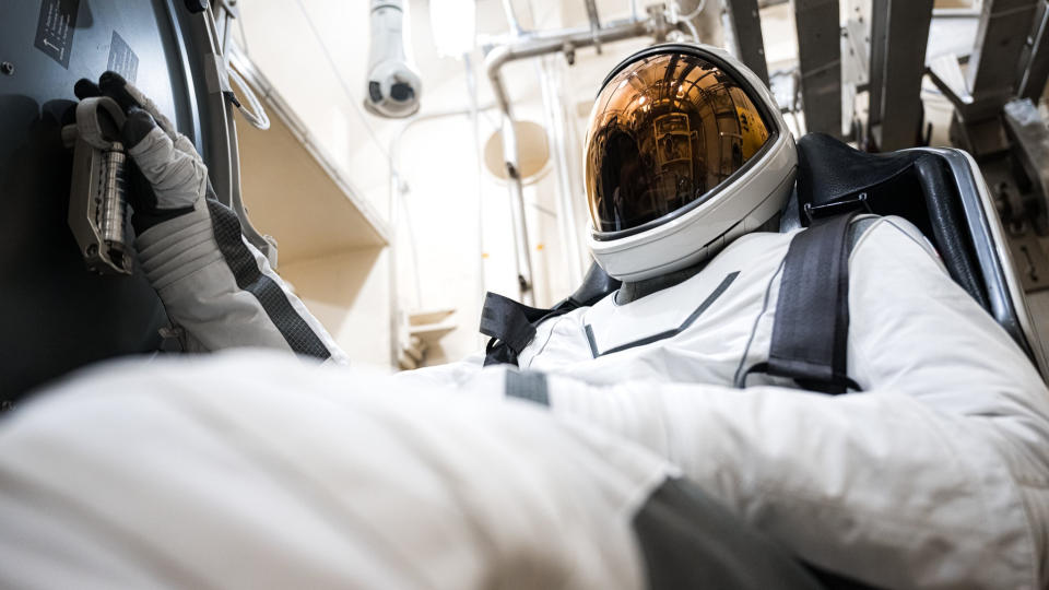  A figure in a clean white spacesuit sits strapped into a black bucket seat, as the image looks upward from his blurry knee on the left in the foreground. Above on the left, a gloved hand with black-tipped fingers holds a component against the dark wall of a spacecraft. The full face visor on the helmet near the top center right reflects a copper shine. Behind, white walls and piping. 