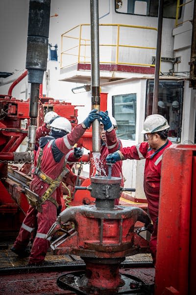 people in protective gear and hard hats on a ship's deck, raising a long metal tube with water coming out the bottom