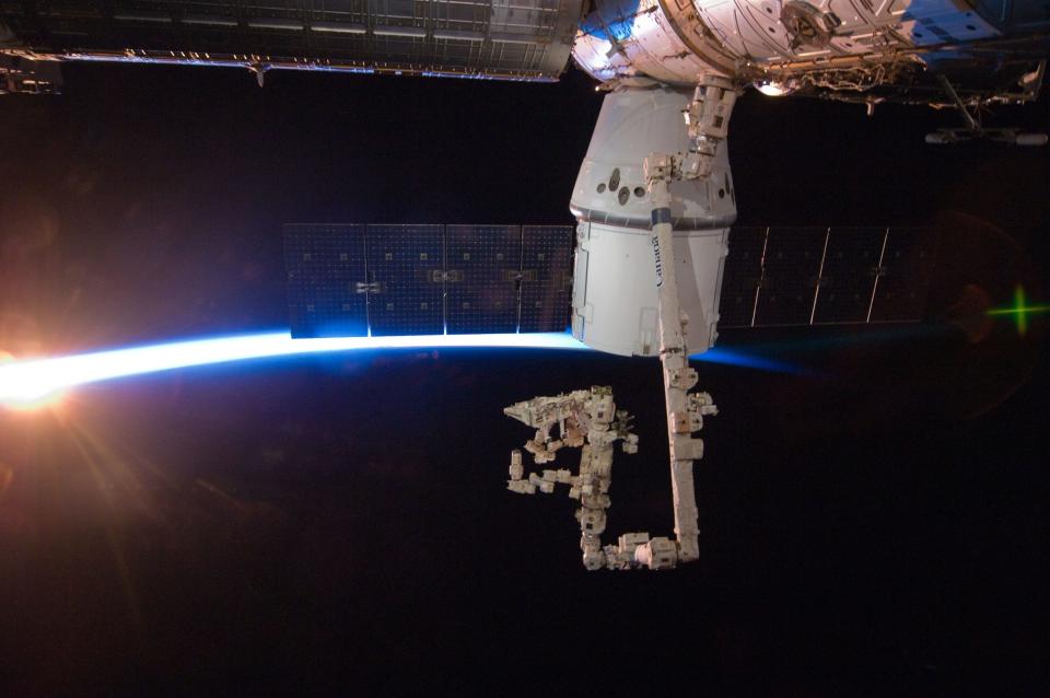 white spacex dragon spaceship docked to a robotic arm of the space station above dark black nighttime earth with a bright blue line on the horizon where sunrays are peeking out for sunrise