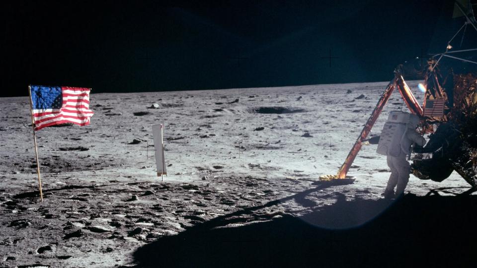  An astronaut in a spacesuit with a gold face shield walks on the grey dusty surface of the moon. 