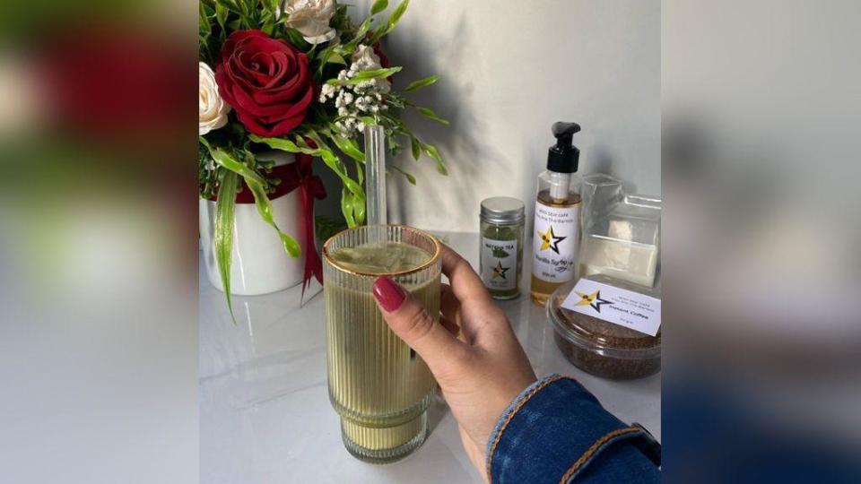 A picture taken from Instagram showing a colourfully-manicured hand holding an iced coffee in front of a bouquet of roses. Plastic pots containing coffee flavourings which all have a star logo are also in the background.