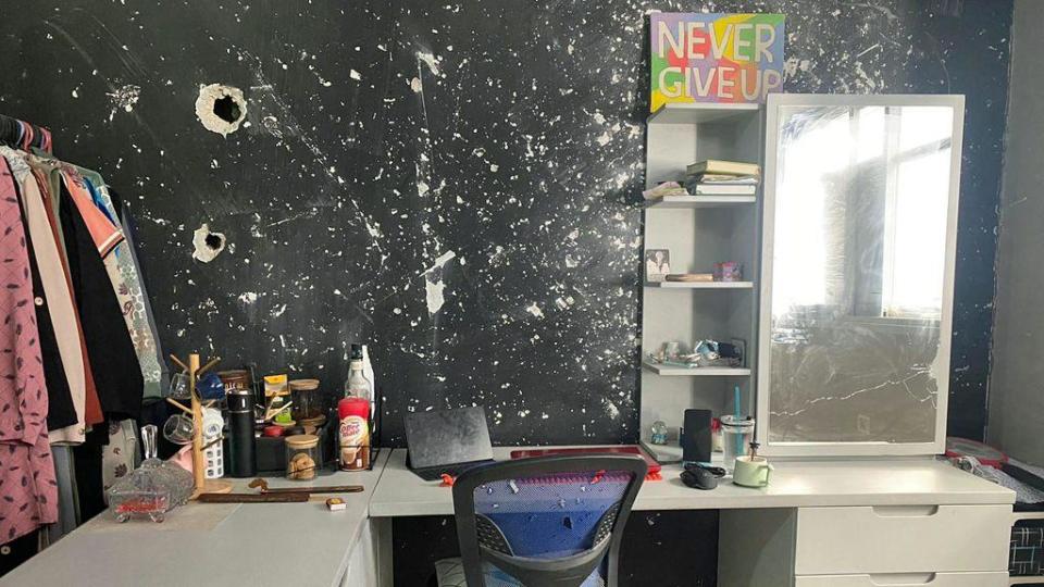 Picture of Asmaa's bedroom - a white desk and computer chair with coffee-related items are neatly on top. The black-painted wall is spattered with white holes and chips from bomb damage. There is a rail of clothes to the left of the photo and a poster which says 'never give up' in white letters on a colourful background on top of a shelf.