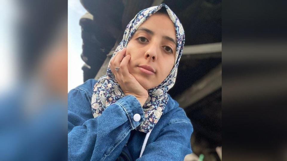 Selfie of Asmaa, whose head rests in her other hand. She has a serious expression on her face. She is wearing a blue denim jacket and a headscarf patterned with blue and white flowers. There appears to be a damaged building behind her