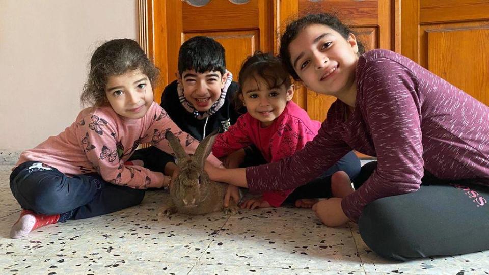 Four children - three girls and a boy - sitting next to each other crossed legged, all leaning forward and smiling, petting a plump rabbit.