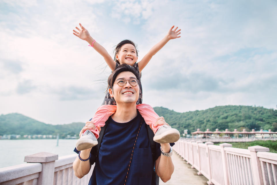 Lovely little girl raising her arms, looking up to the sky joyfully while her dad carrying her on shoulders in promenade.