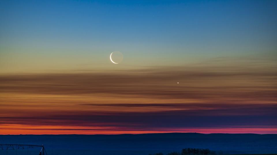 The conjunction of the waning crescent moon with Venus as they were rising low in the northeast dawn sky in southern Alberta, Canada. Earthshine is visible on the dark side of the moon. The sky exhibits the wonderful transition of colours from the orange at the horizon through the spectrum to the blues at top