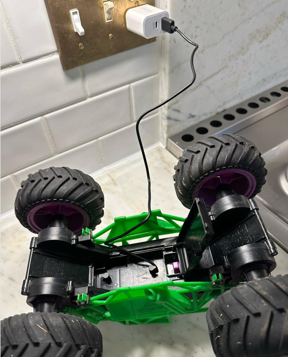 A green and black toy monster truck on a countertop, plugged into a charger on the wall.