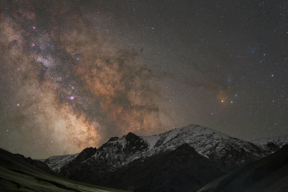 At the base of the image are towering snow-capped mountains and above the Milky Way dense with stars rises up into the sky.