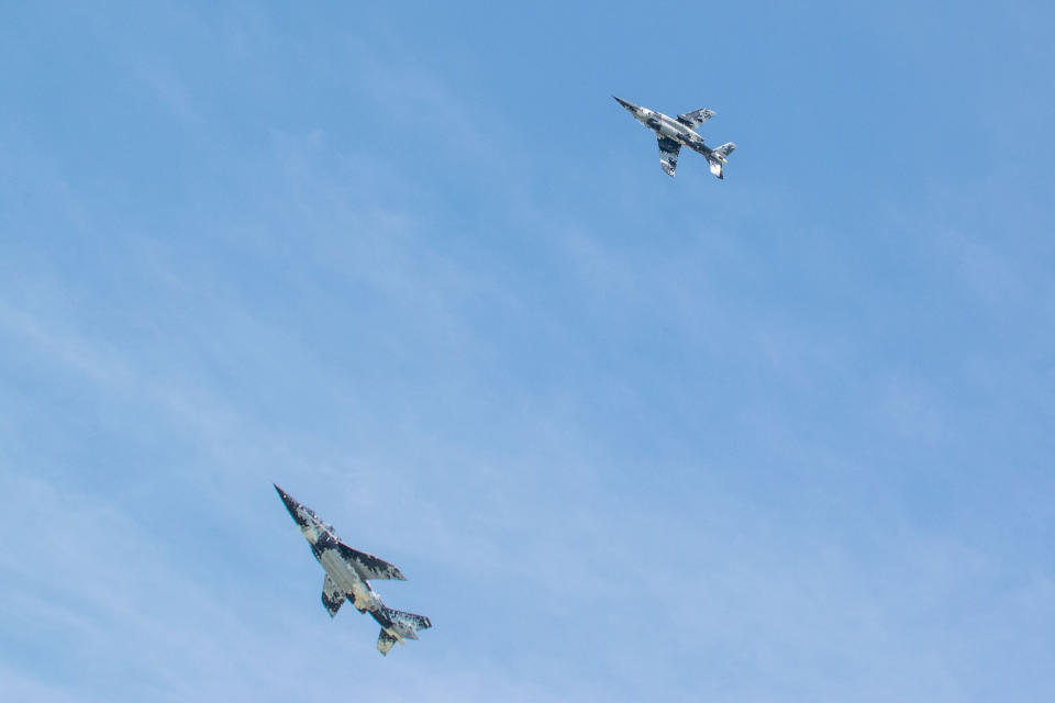 Two blue and white camo jets fly through the blue sky.