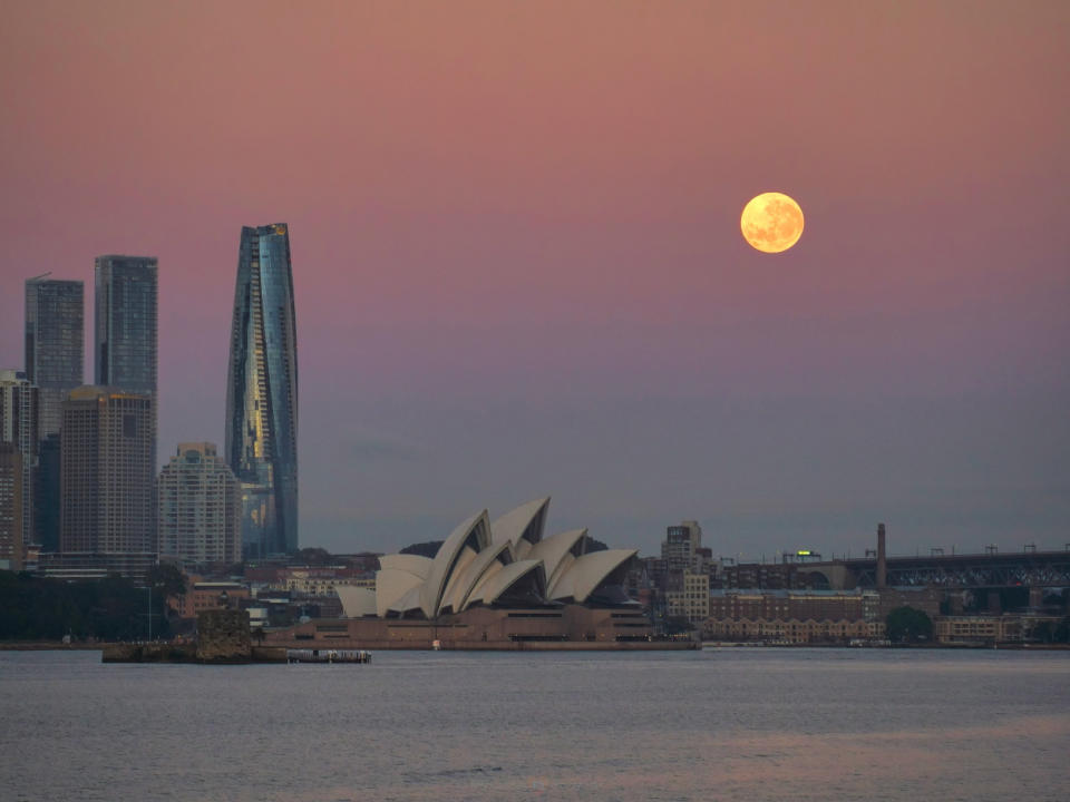 a dusty pink sky with a yellow hued full moon in the top right corner of the image with the Sydney skyline below, including the Sydney Opera House.