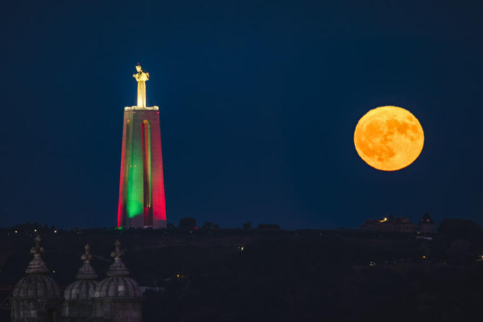 a large statue of Christ on the left is illuminated with green and red lights while the full moon shines brightly in the sky to the right of the statue.