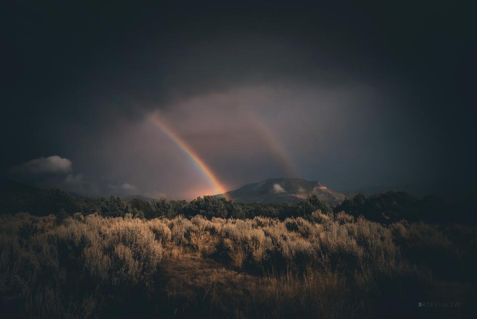 a faint but colorful double rainbow rises out from the landscape into a cloudy sky.