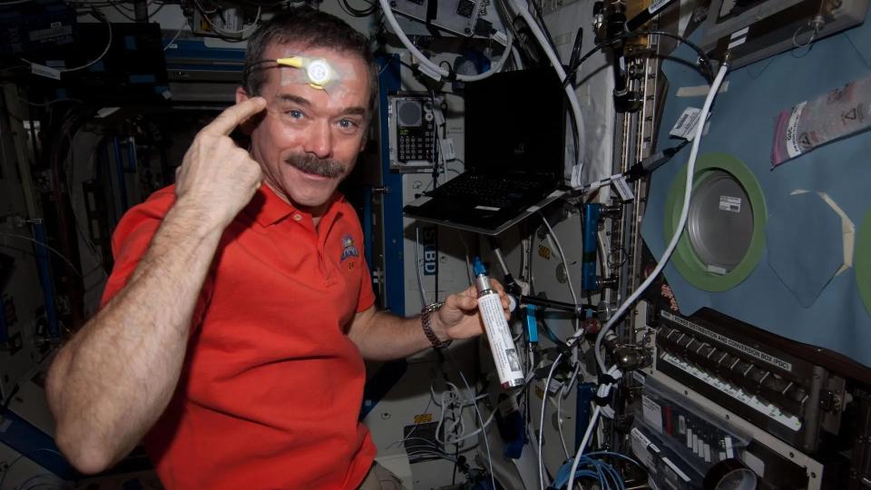 an astronaut pointing at a skin sensor on his forehead while floating in a space station module