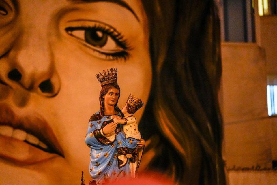 An effigy of the Virgin Mary is carried above the heads of people (who are out of eyeshot). In the background, there is a large mural of a woman's face. 