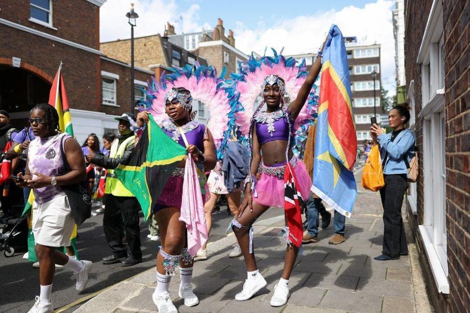 Shauna-Leigh, 11, holds a flag of Jamaica and La'mya, 13, holds a flag of the Democratic Republic of Congo as they take part in the Children's Day Parade at Notting Hill Carnival. They are pictured posing with their flags on the street, as other people pass by. They are wearing white trainers, pink skirts, purple tops with a jewelled necklace overlaying them, and pink and blue feathered costume pieces that resemble halos around their heads.