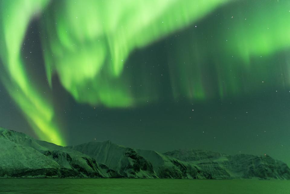 ribbons of green light dance in the sky above snow capped mountains and reflect off the water below.