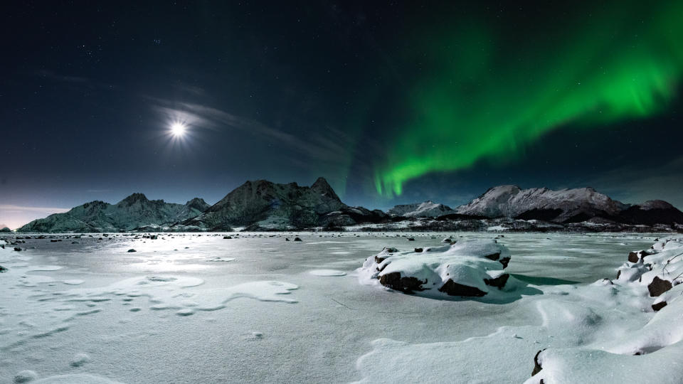 northern lights and a bright full moon illuminate the sky above snowcapped mountains and a frozen body of water.